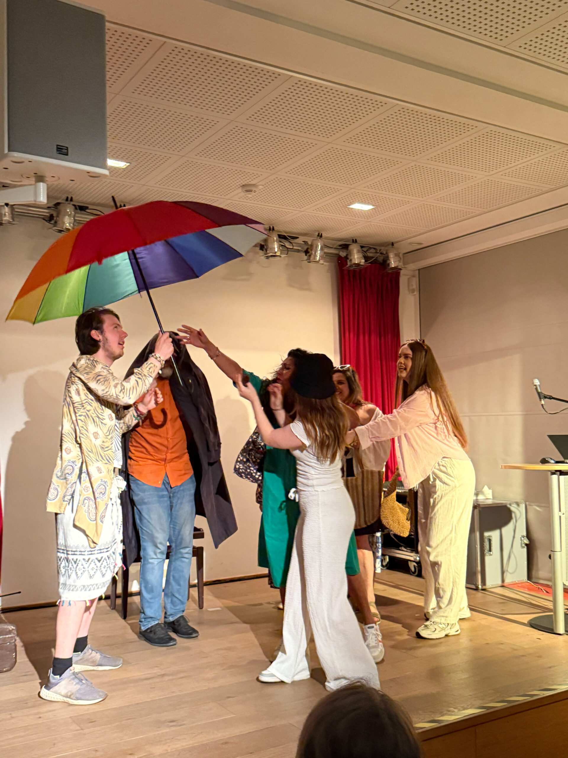 Das Foto zeigt eine Schüler:innen-Gruppe auf einer kleinen Bühne, sie streiten sich um einen regenbogenfarbenen großen Regenschirm